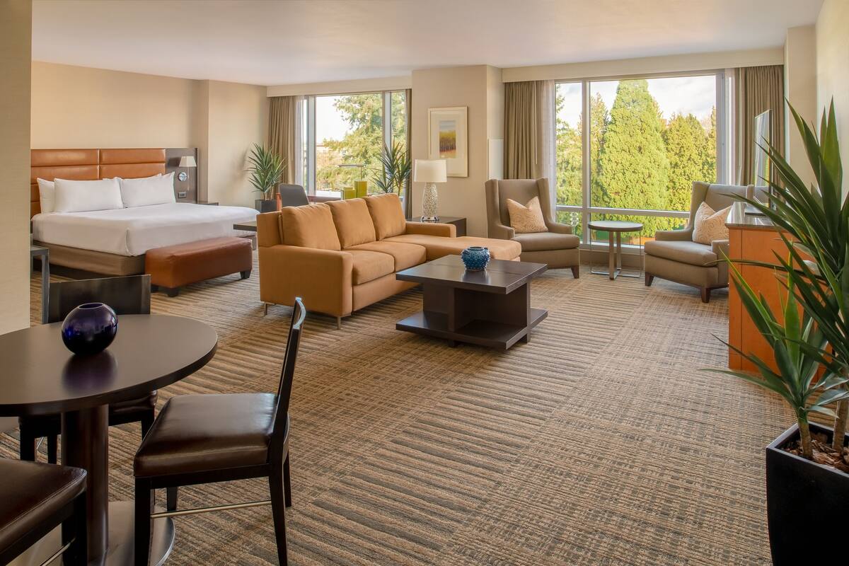 A hotel room with a bed, a sofa, a coffee table, a dining table with chairs, and a small balcony with a view of trees.