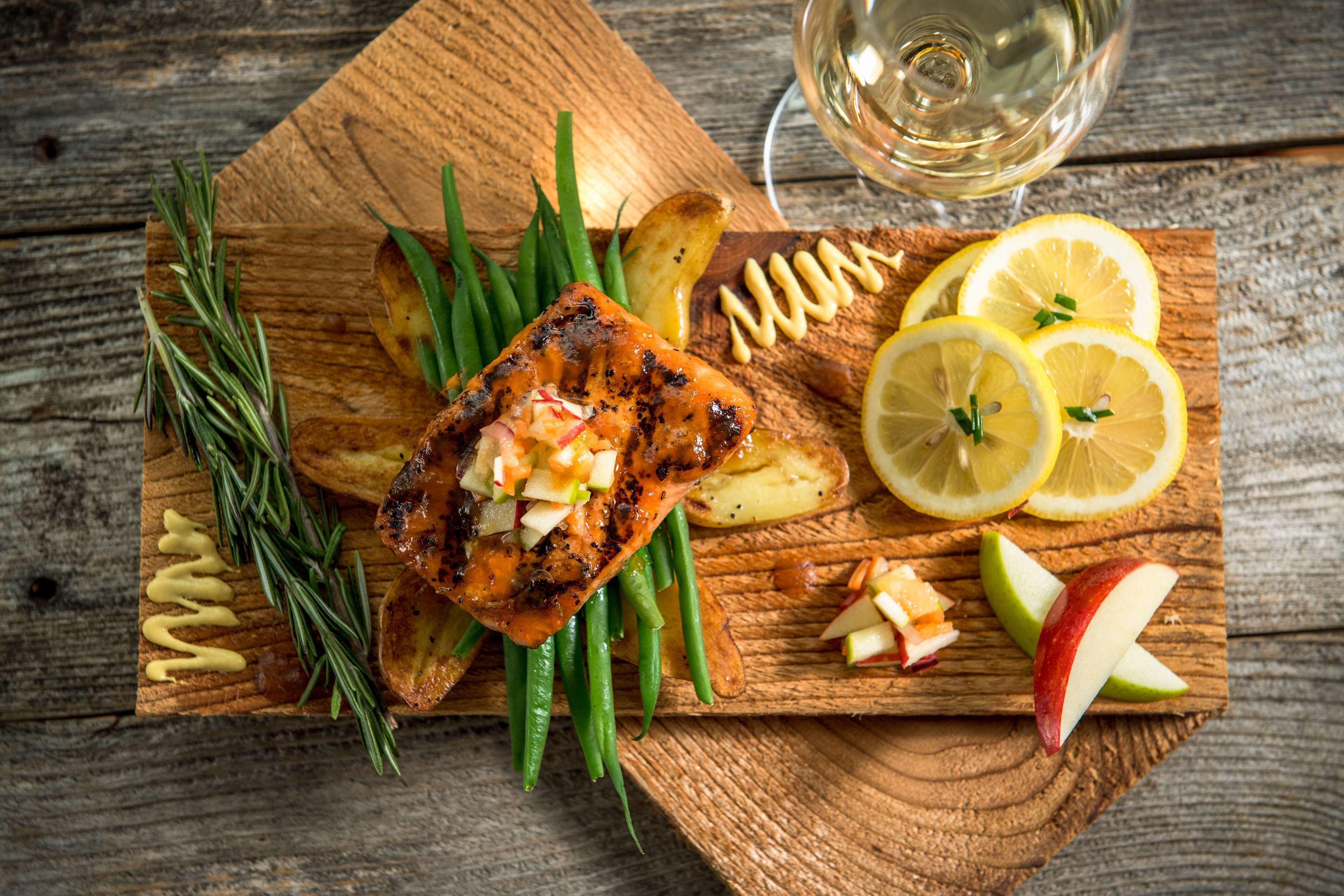 Ein Holzbrett mit gegrilltem Lachs, grünen Bohnen, Kartoffeln, Zitronenscheiben und Apfelscheiben.