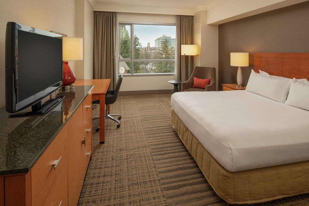 A hotel room with a bed, a desk with a TV, a chair, a small table, and a window with curtains.