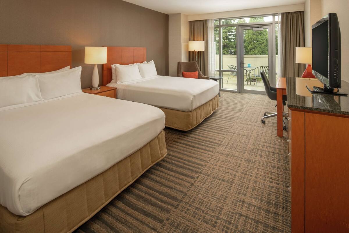 A hotel room with two beds, a desk with a TV, and a balcony with a view.
