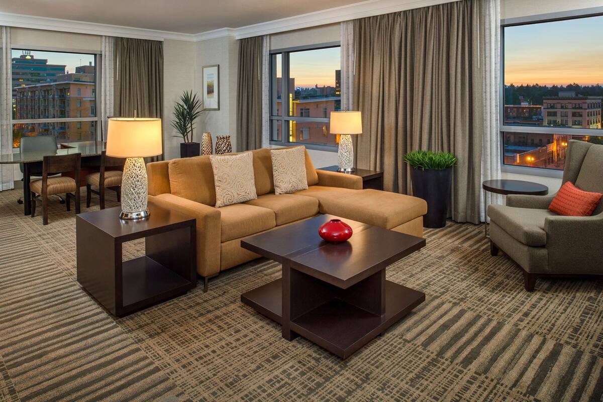 A hotel room with a sofa, two armchairs, a coffee table, a side table, a lamp, a plant, and a view of the cityscape through large windows.