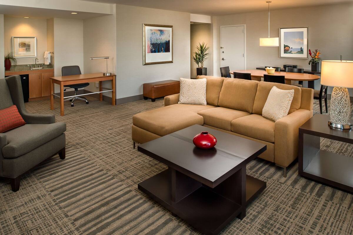A hotel room with a sofa, armchair, dining table, and a desk with a chair.