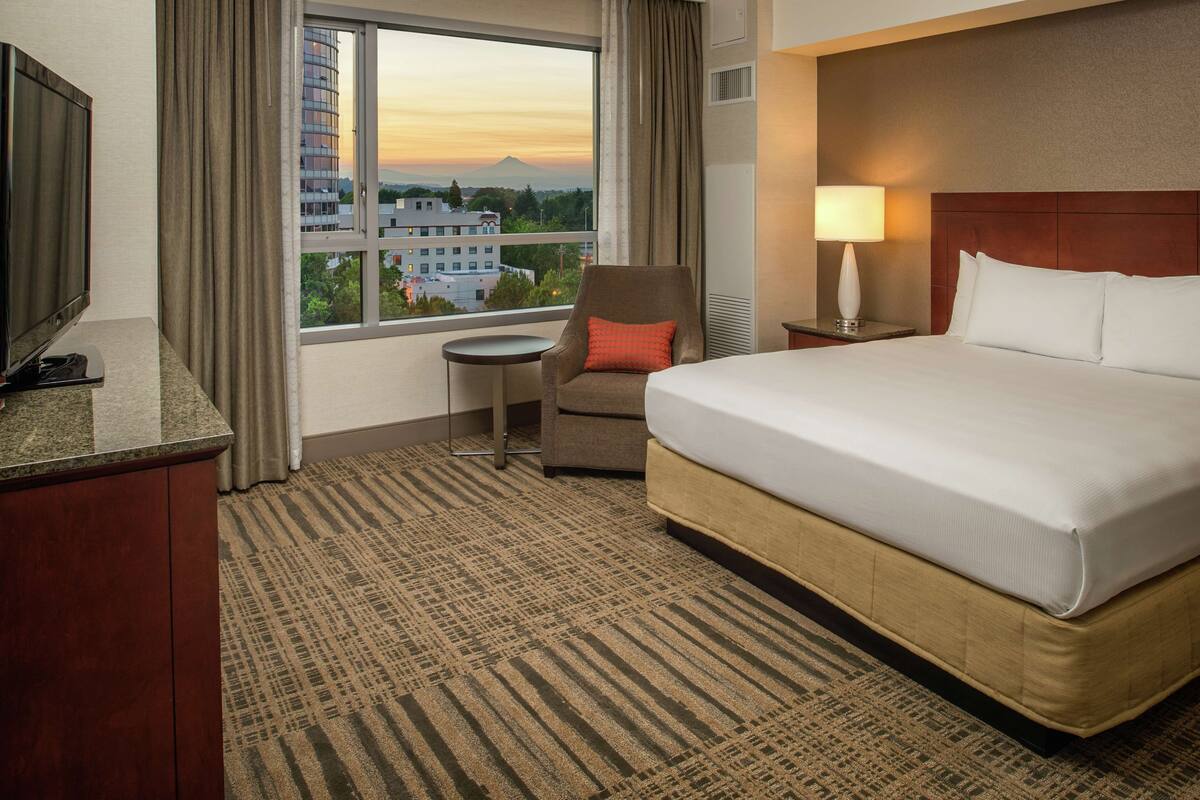 A hotel room with a bed, a chair, a small table, a TV, and a window with a view of buildings and a mountain.
