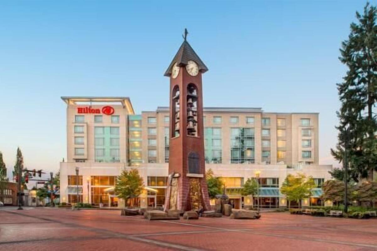 A hotel with a clock tower, a large building with a Hilton sign, and a paved plaza with trees.