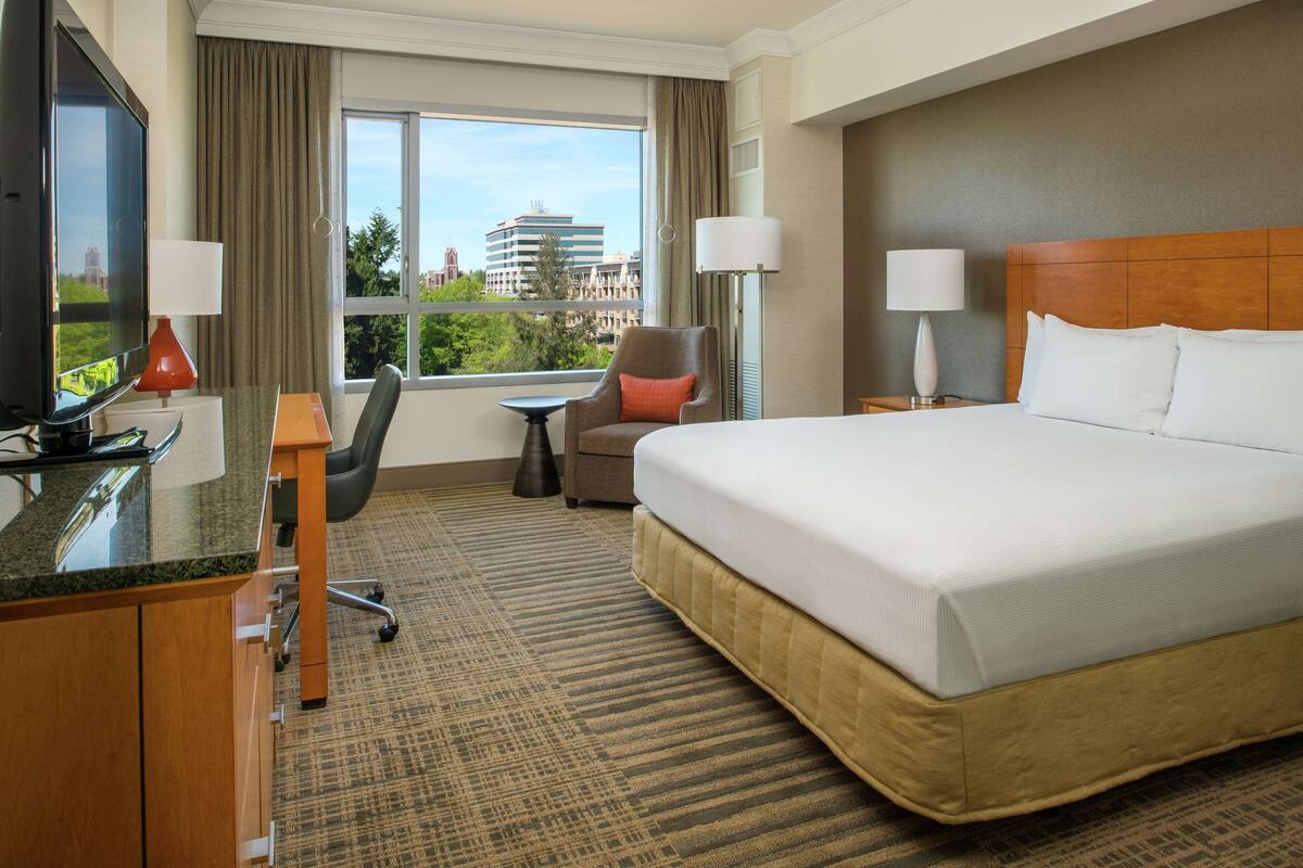 A hotel room with a large bed, a desk with a TV, a chair, and a view of a cityscape through the window.