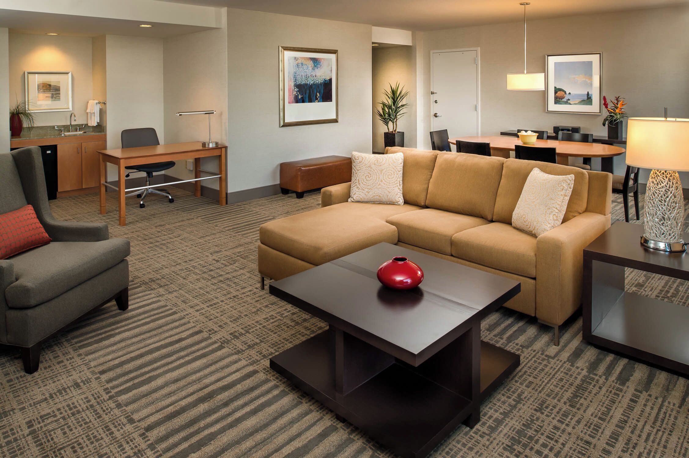 A hotel room with a sofa, armchair, dining table, and a desk with a chair.