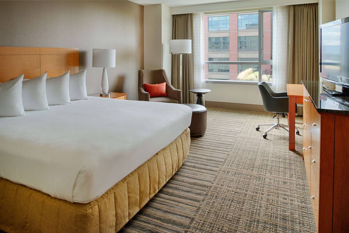 A hotel room with a large bed, a desk, a chair, a TV, and a window with a view of buildings.