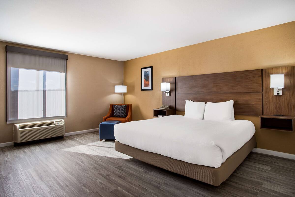 A hotel room with a large bed, a chair, a desk, a lamp, and a window with blinds.