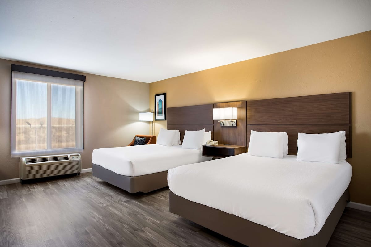 A hotel room with two beds, a large window, a wall-mounted heater, and a nightstand with a lamp.