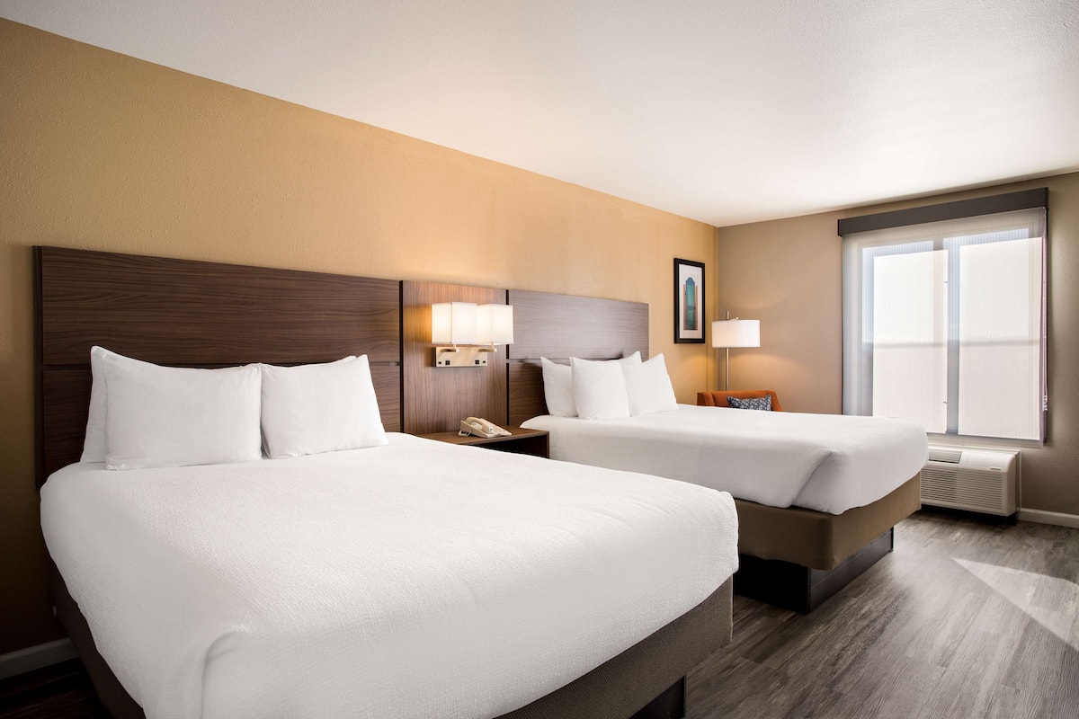 A hotel room with two beds, a nightstand with a phone, a wall-mounted lamp, and a large window with blinds.