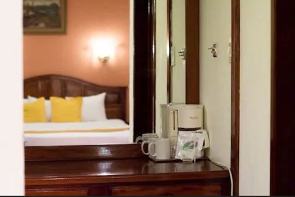 Una habitación de hotel con cama, una mesita de noche con cafetera y taza, y una puerta con cerradura.