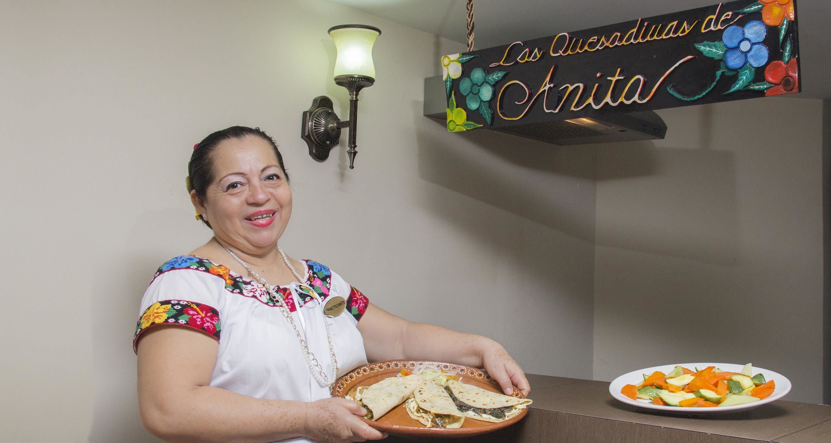 Une personne tenant une assiette de nourriture, debout devant un mur sur lequel est affiché un panneau indiquant : « Les quesadillas d’Anita ».