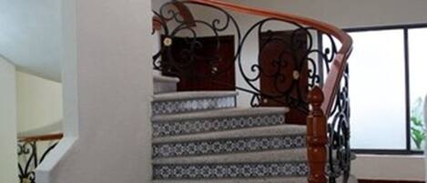 Uma escadaria com degraus estampados e um corrimão de madeira, conduzindo a um patamar com guarda-corpo metálico curvo.