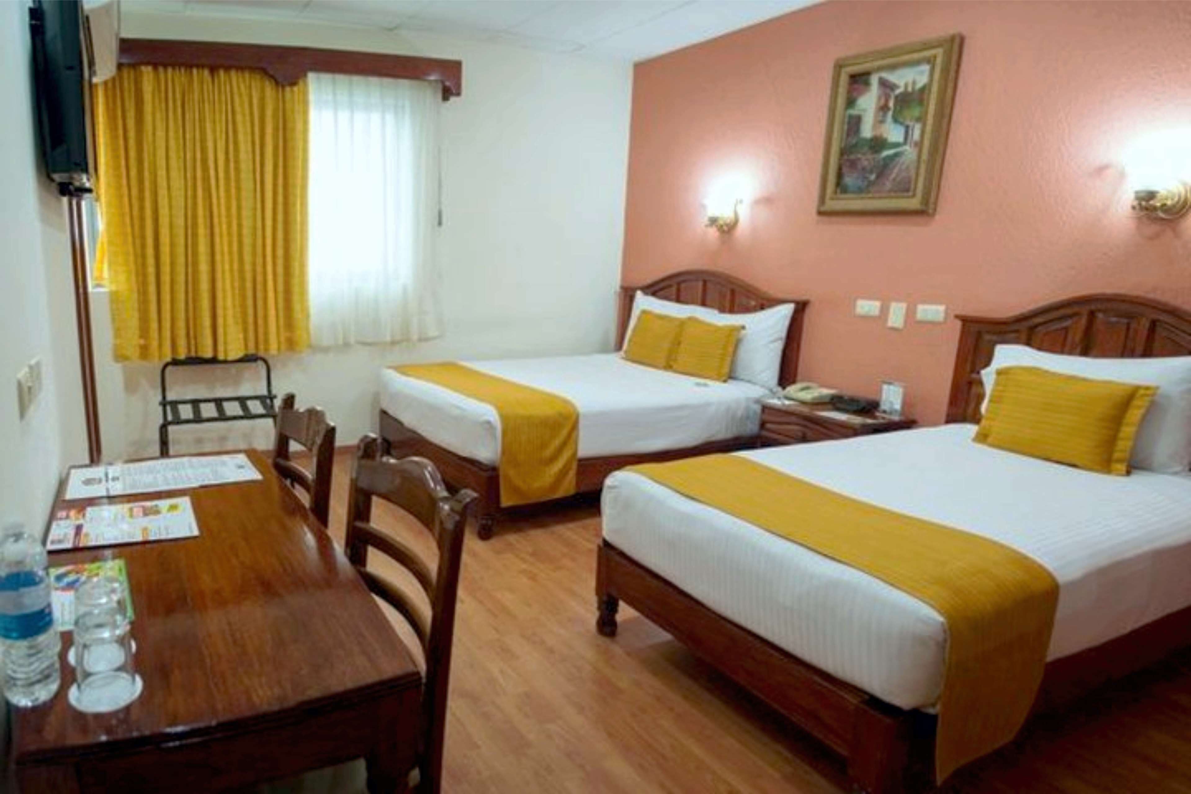 A hotel room with two beds, wooden furniture, a table with a chair, a window with yellow curtains, and a painting on the wall.