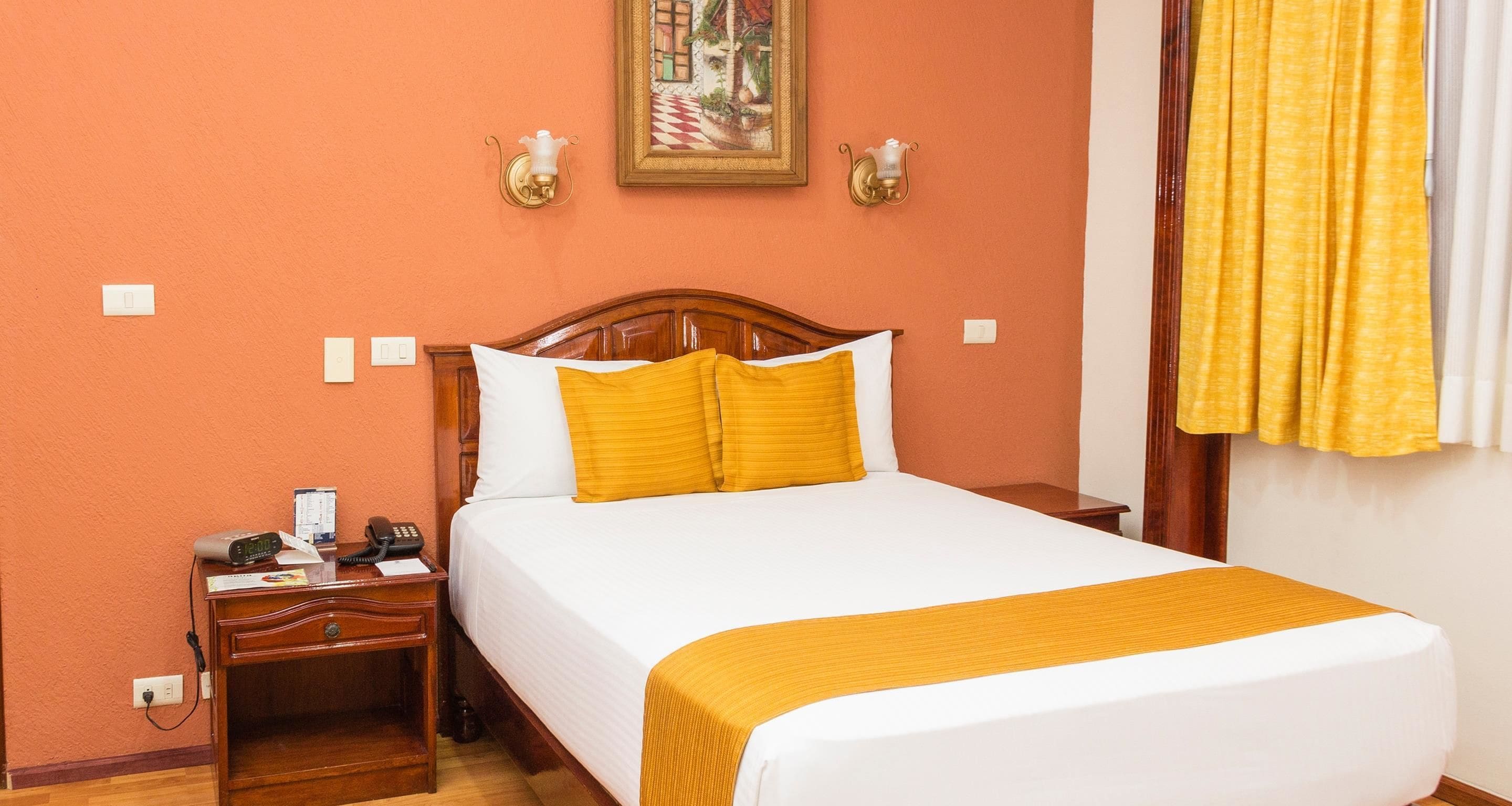 A hotel room with a wooden bed, white bedding, and a yellow throw. There is a nightstand with a phone and a lamp, and a painting on the wall.