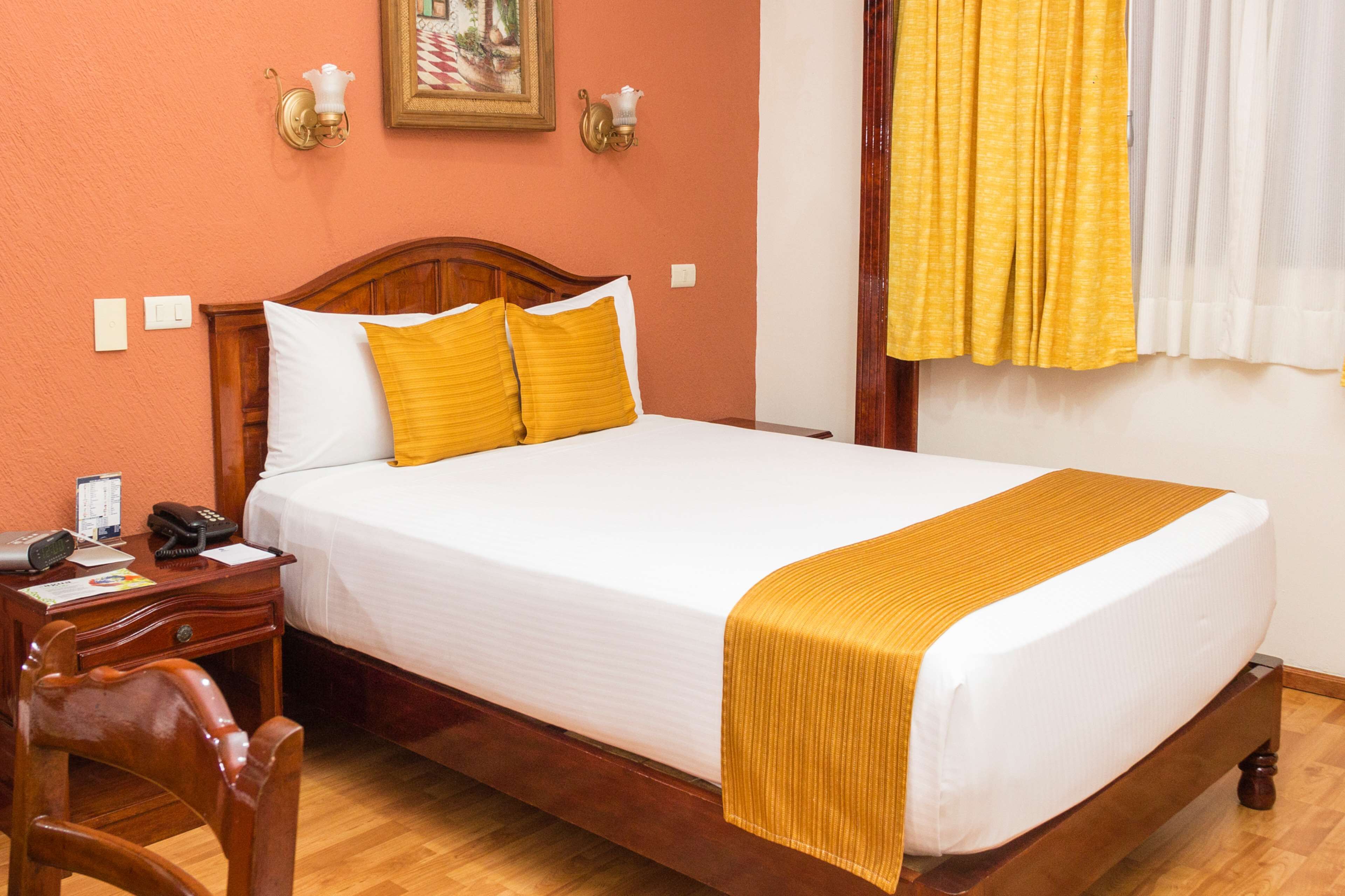 A neatly made bed with white and yellow bedding, a wooden headboard, and a bedside table with a lamp and a telephone.