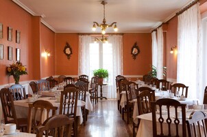 A dining room with multiple tables and chairs, a chandelier, and a large window with curtains.