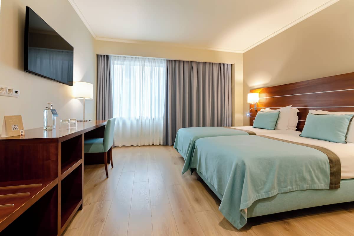 Habitación de hotel con un escritorio de madera, un televisor de pantalla plana, dos camas con ropa de cama azul claro, un ventanal con cortinas y un cabecero de madera.