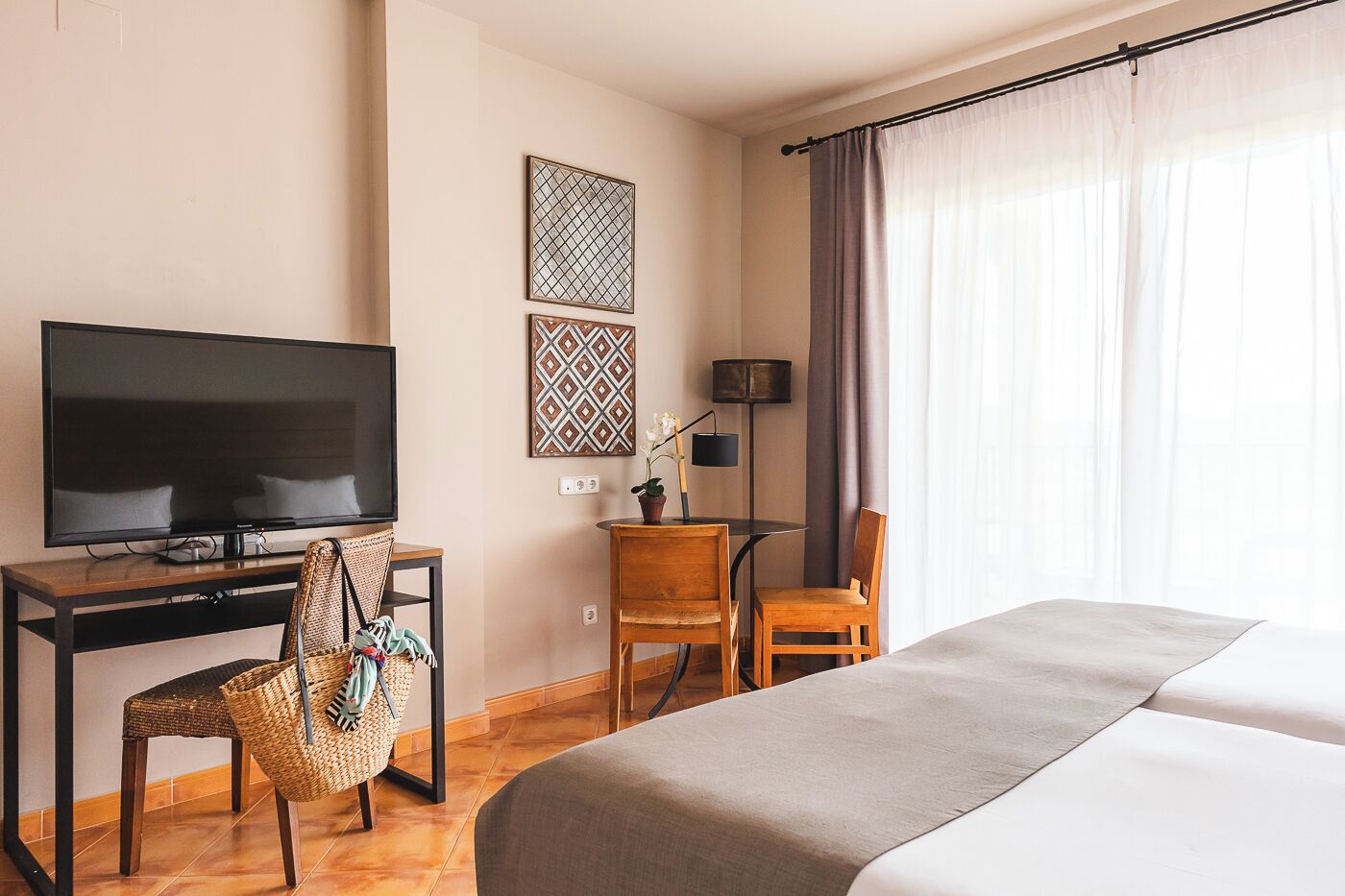 A hotel room with a bed, a desk with a TV, a chair, a lamp, and two decorative wall pieces.