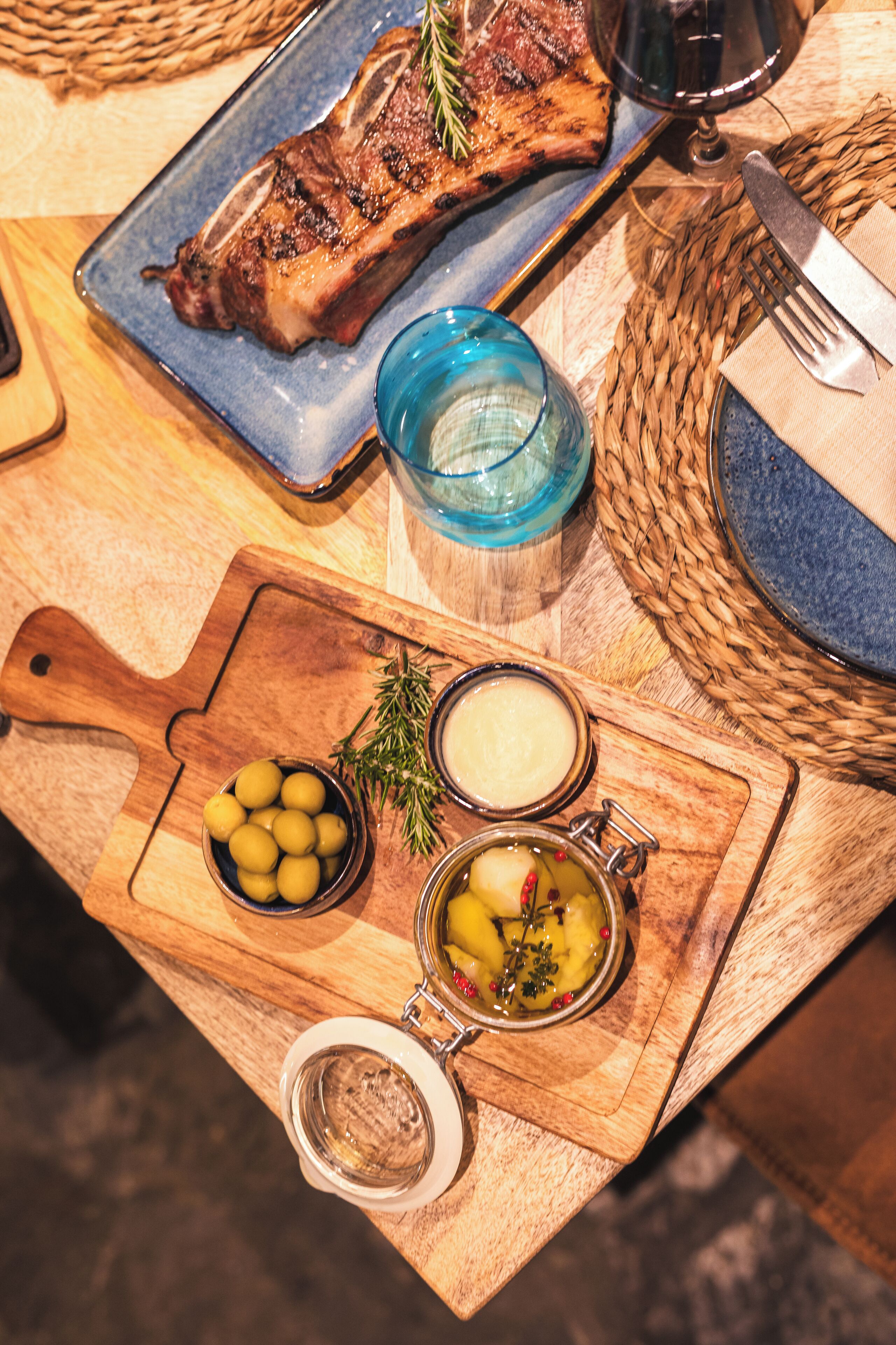 A wooden table with a plate of steak, a bowl of olives, and a jar of pickles.