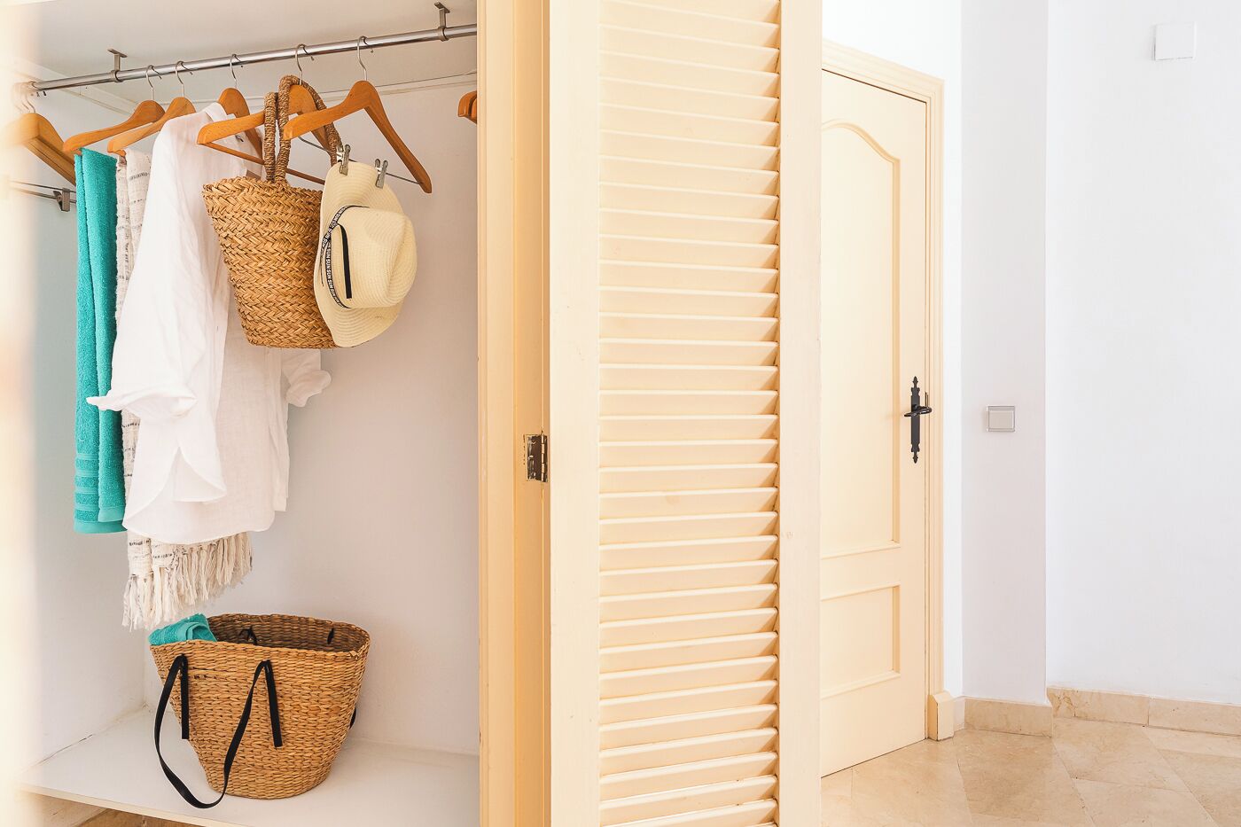 A walk-in closet with a wooden door, a hanging rack with clothes and baskets, and a tiled floor.