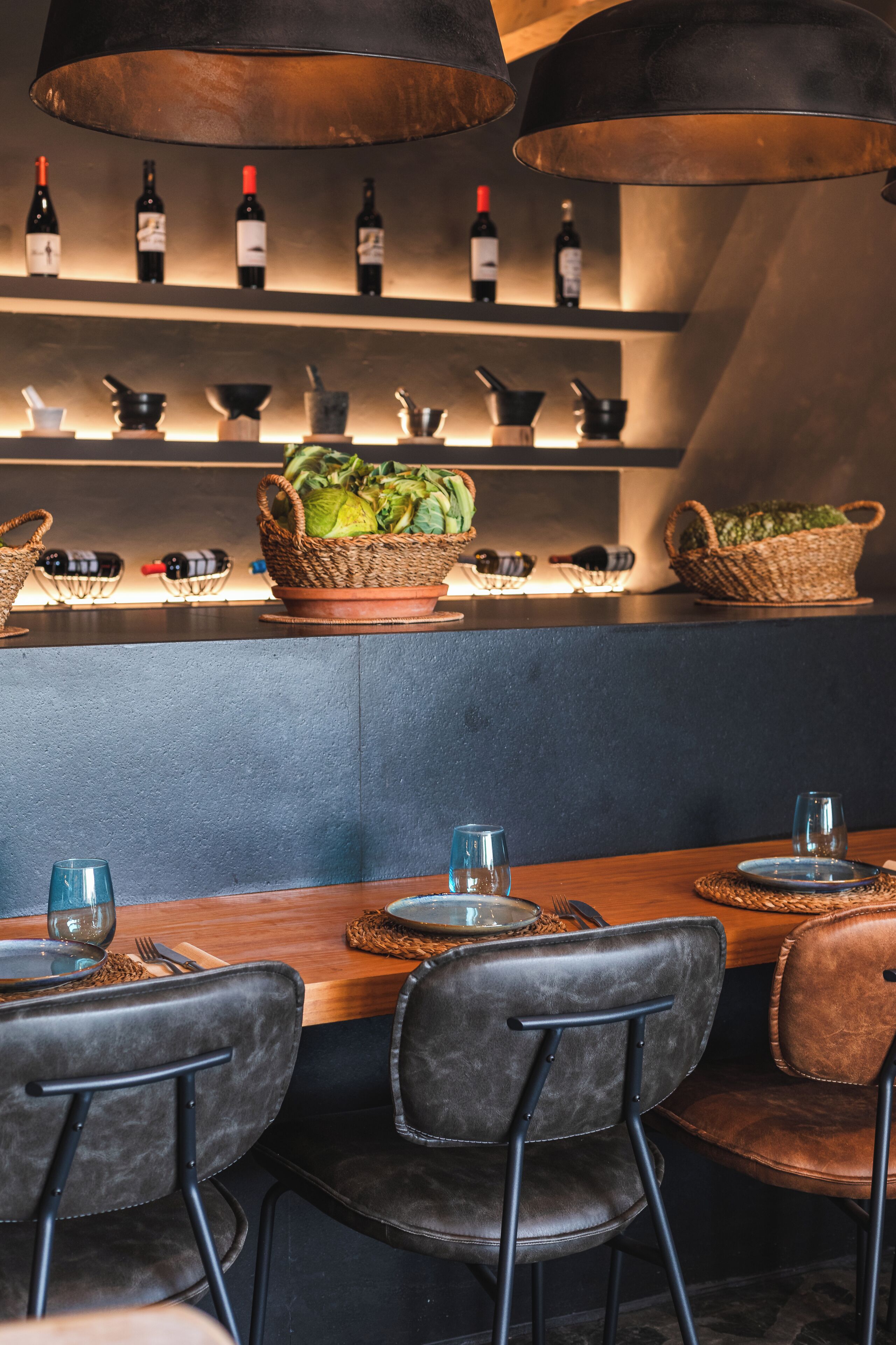 A restaurant interior with a long wooden table, black leather chairs, and a wine display above.