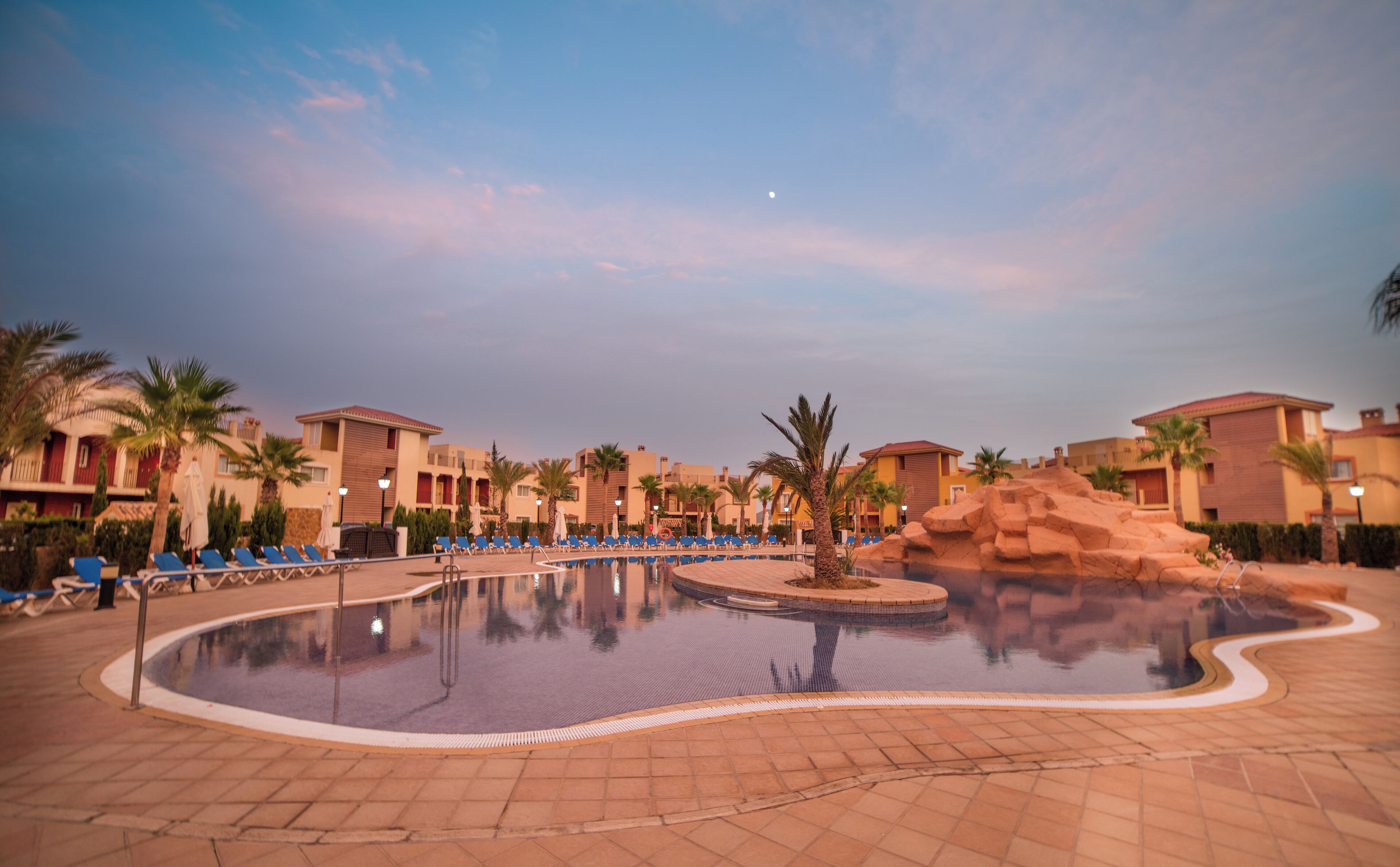 Zona de piscina en un resort con sillones de descanso y palmeras.