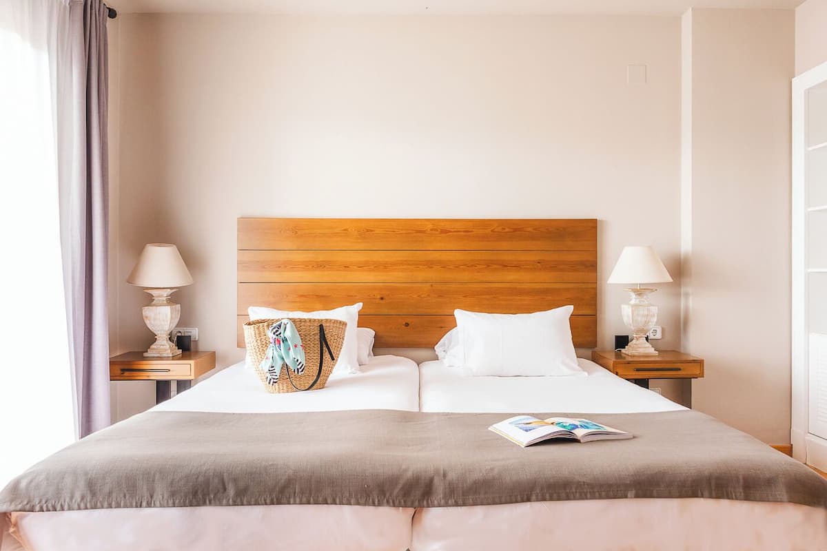 Een netjes opgemaakt bed met wit en grijs beddengoed, een houten hoofdbord en twee nachtkastjes met lampen.