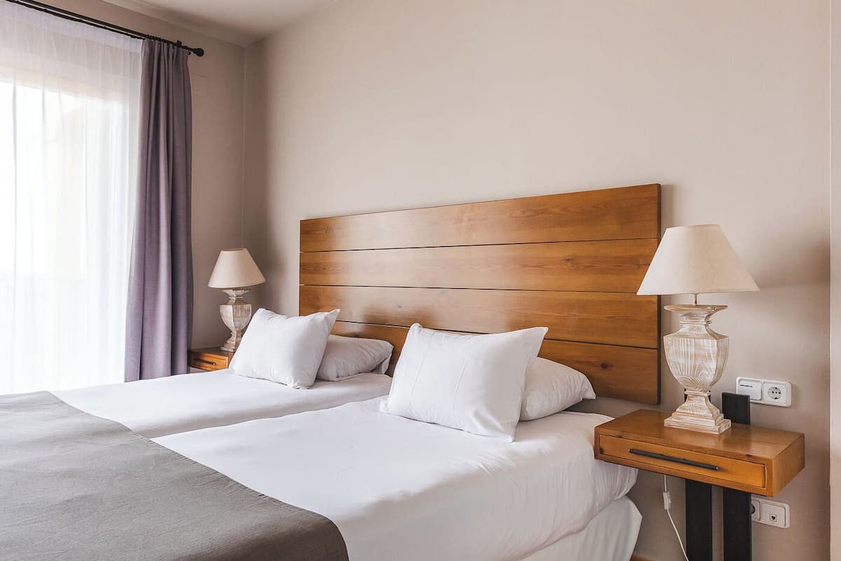 Een hotelkamer met twee bedden, een houten hoofdeinde, nachtlampjes en een raam met gordijnen.