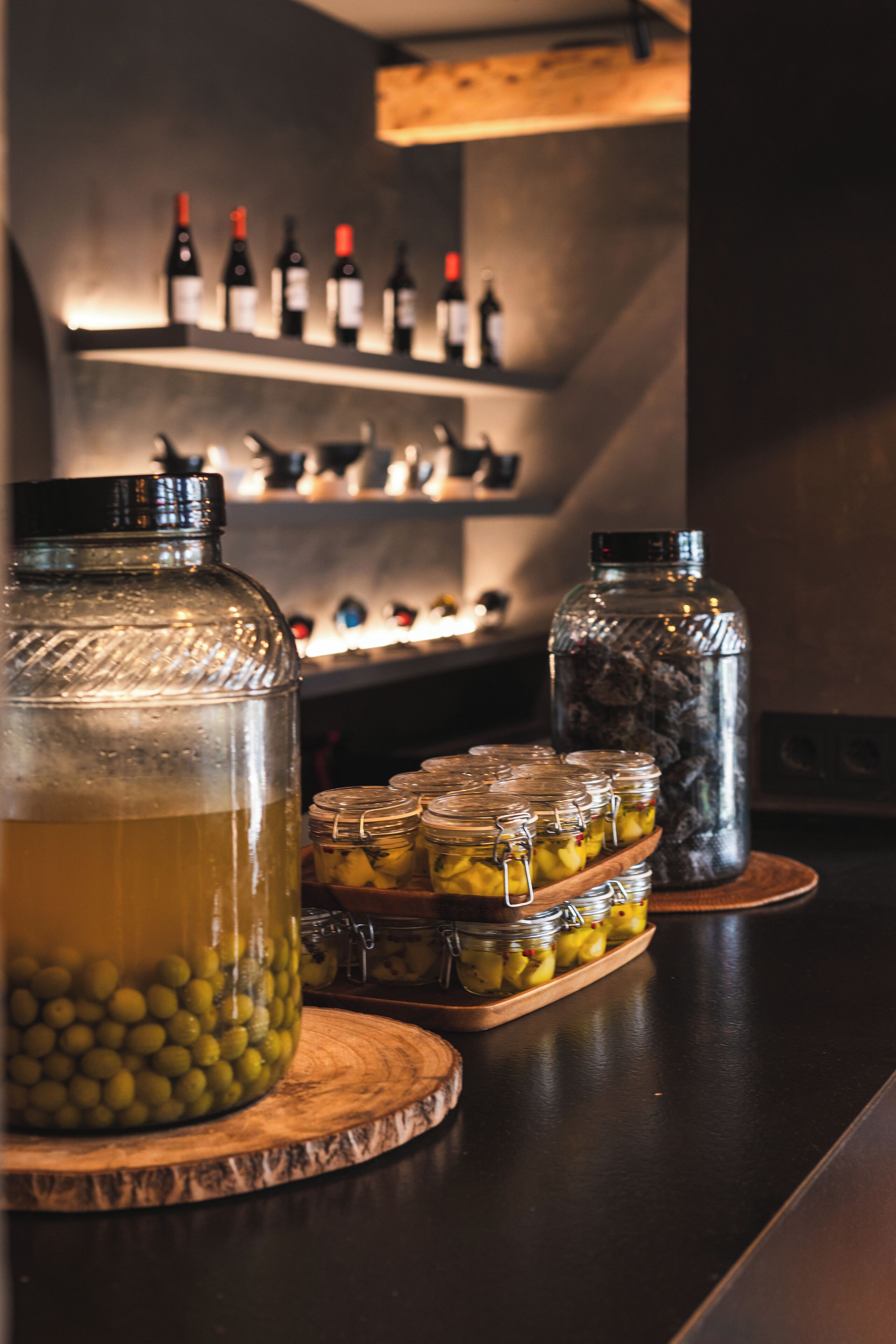 A bar area with a selection of wine bottles, a large glass container with a yellow liquid, and several glass jars with pickles on a wooden board.