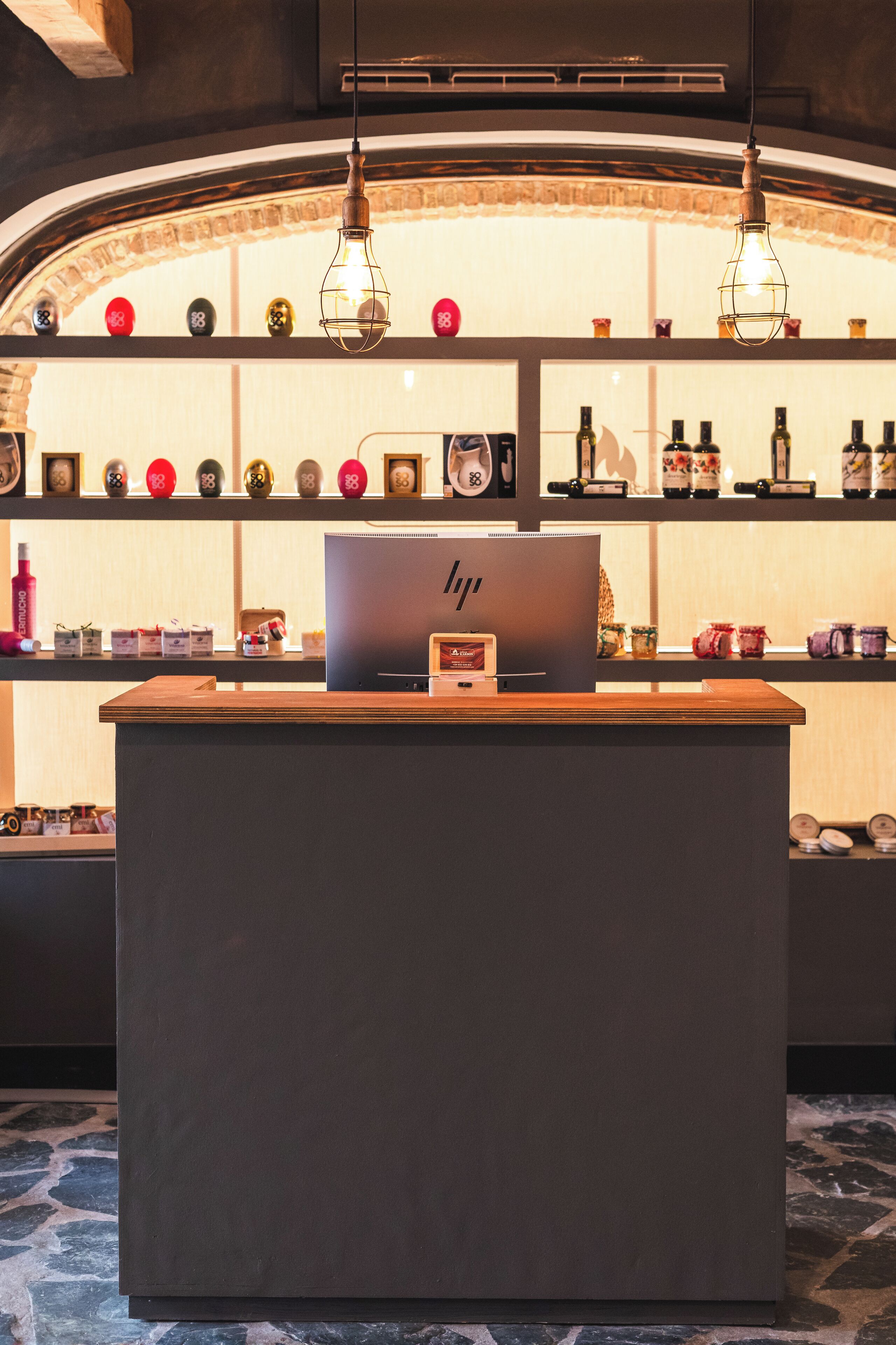 A reception desk with a laptop, shelves with bottles and decorative items, and hanging light fixtures.