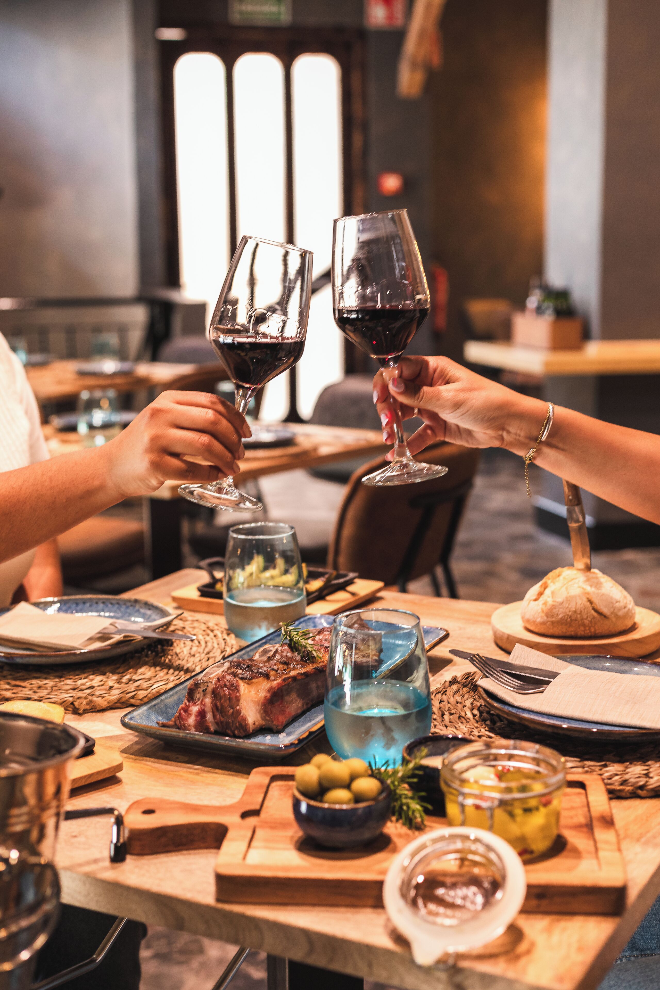 Uma mesa de jantar com carne assada, pão, azeitonas e um copo de vinho tinto.