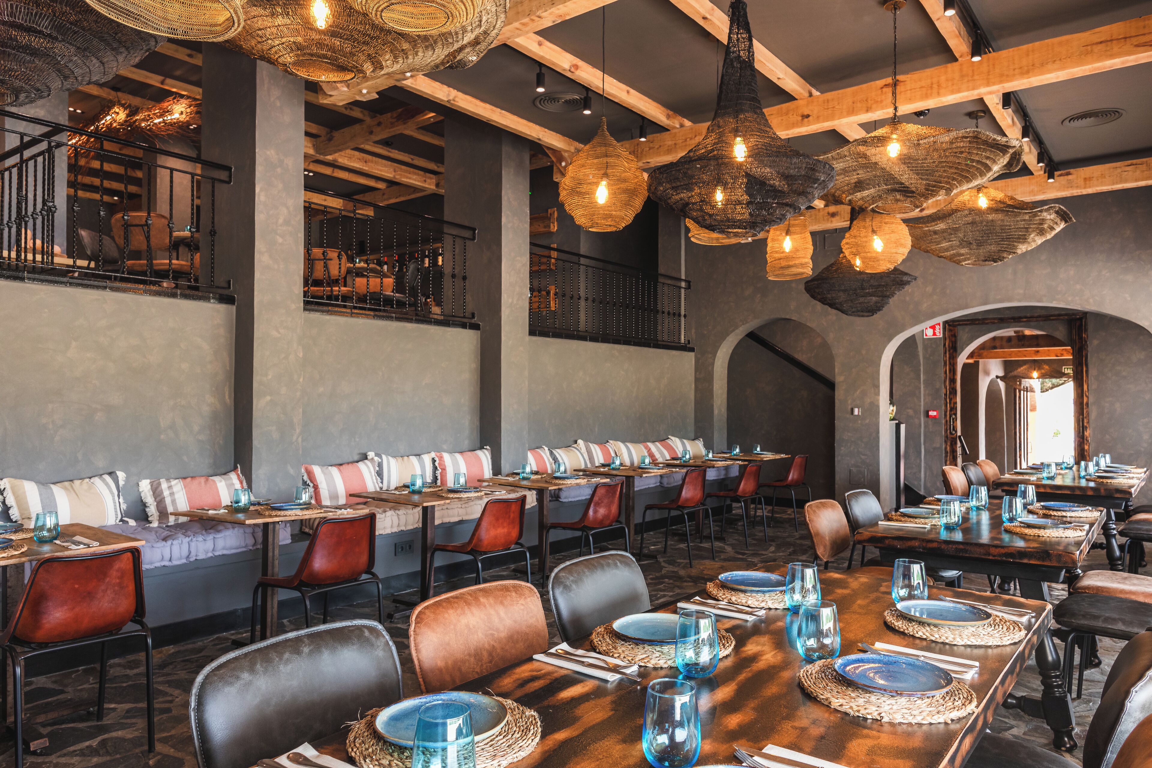 A restaurant with long communal tables, cushioned seating, and hanging woven light fixtures.