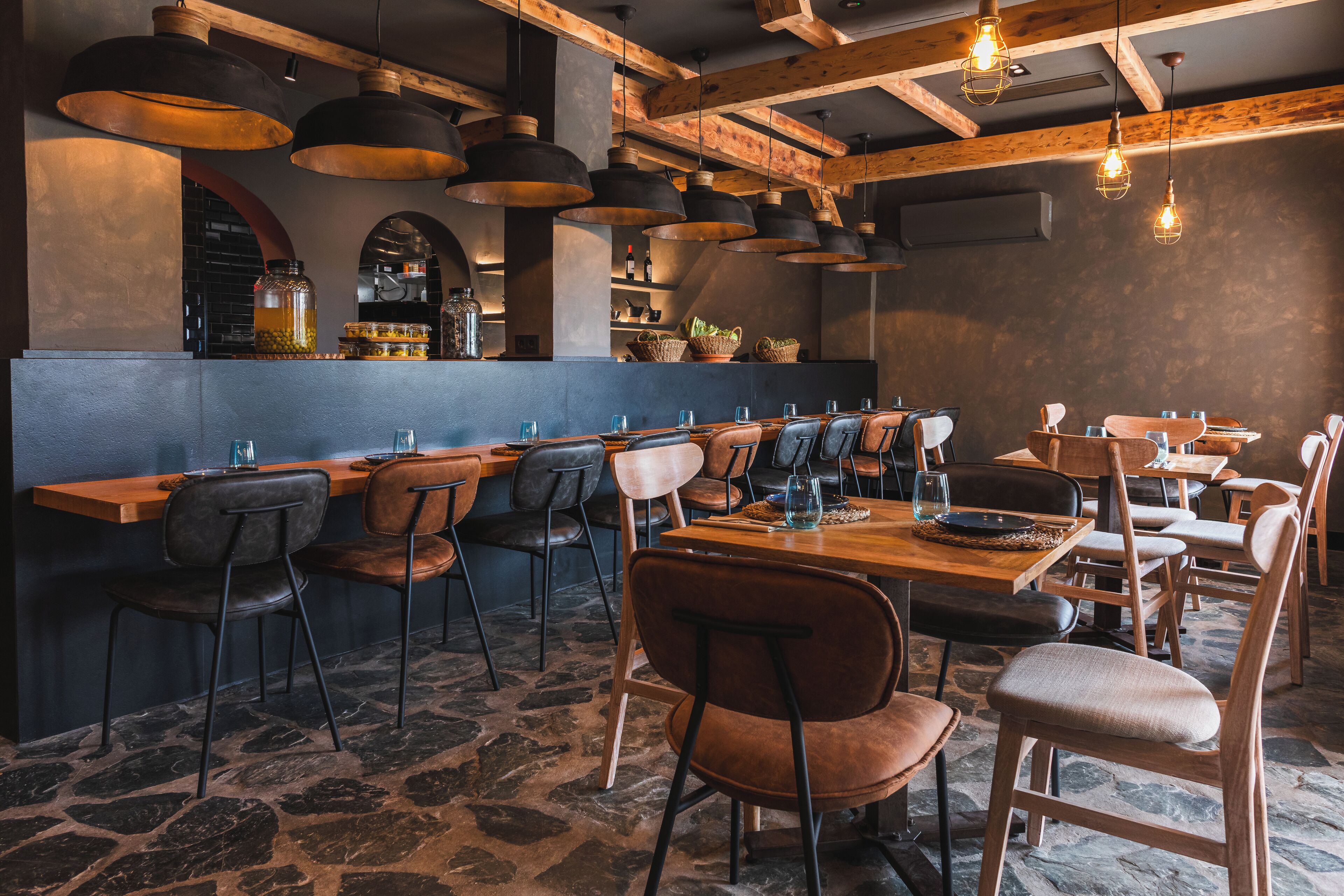 Un restaurante con una barra larga, varias mesas y sillas. Hay luces colgantes y suelo de piedra.