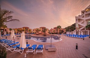 A resort pool area with blue lounge chairs and umbrellas, surrounded by palm trees and buildings.