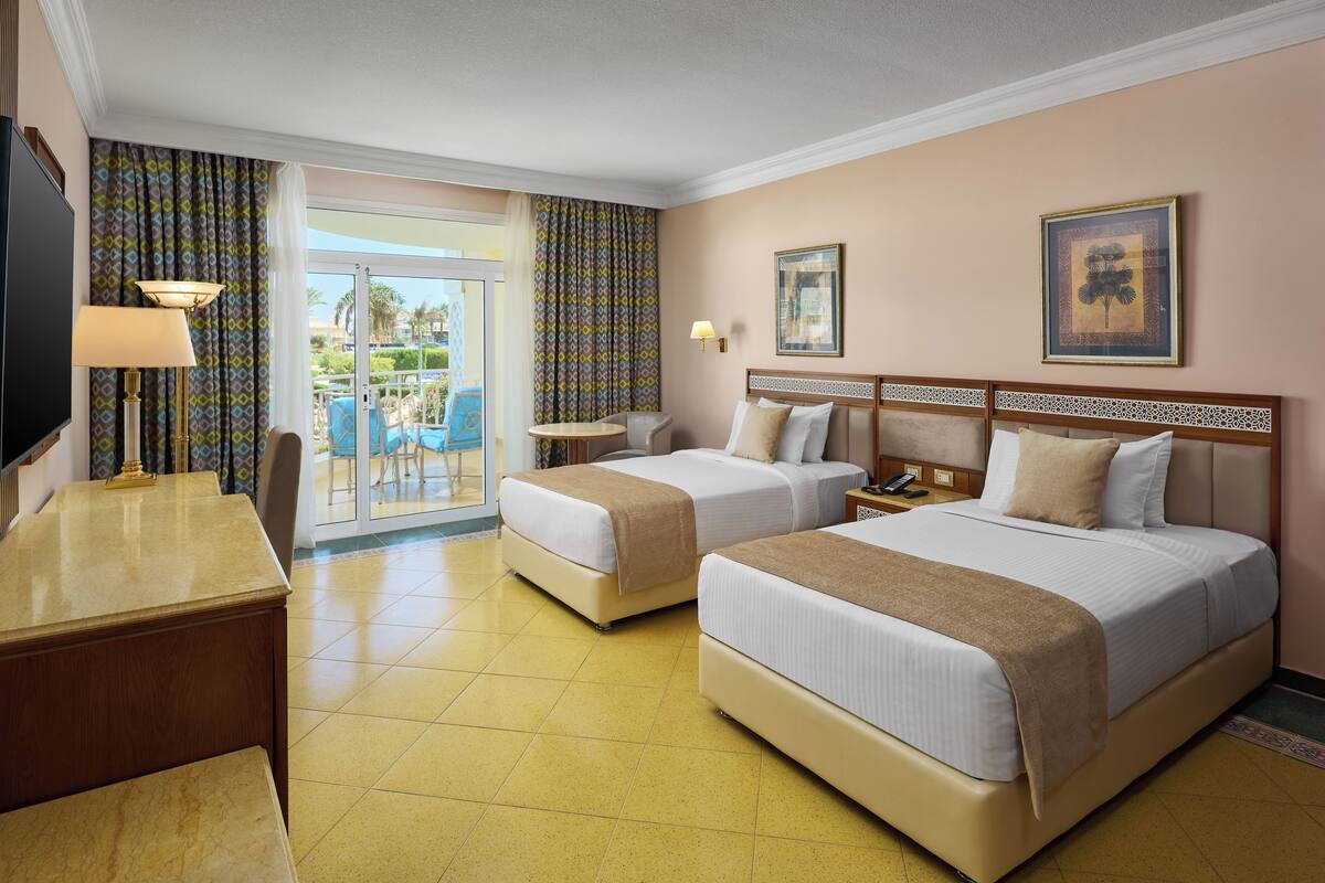 A hotel room with two beds, a desk, a chair, a small table, and a balcony with a view.