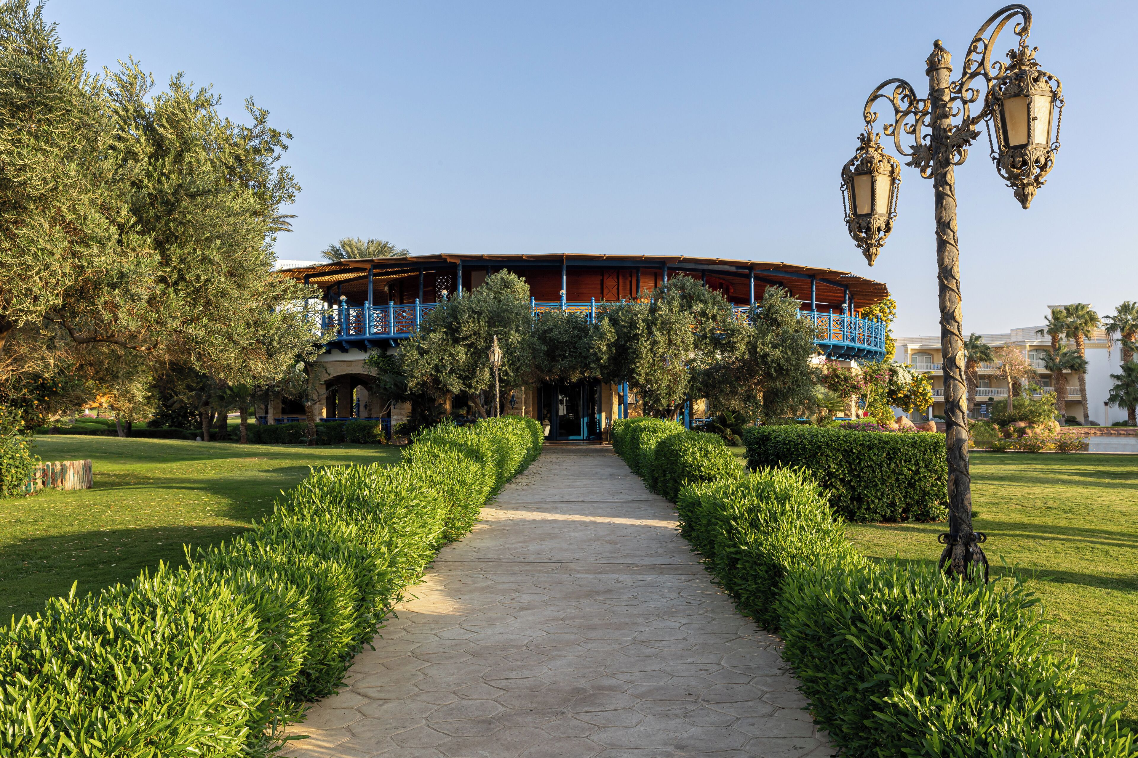 A pathway leading to a building with a blue balcony, surrounded by well-maintained hedges and trees.
