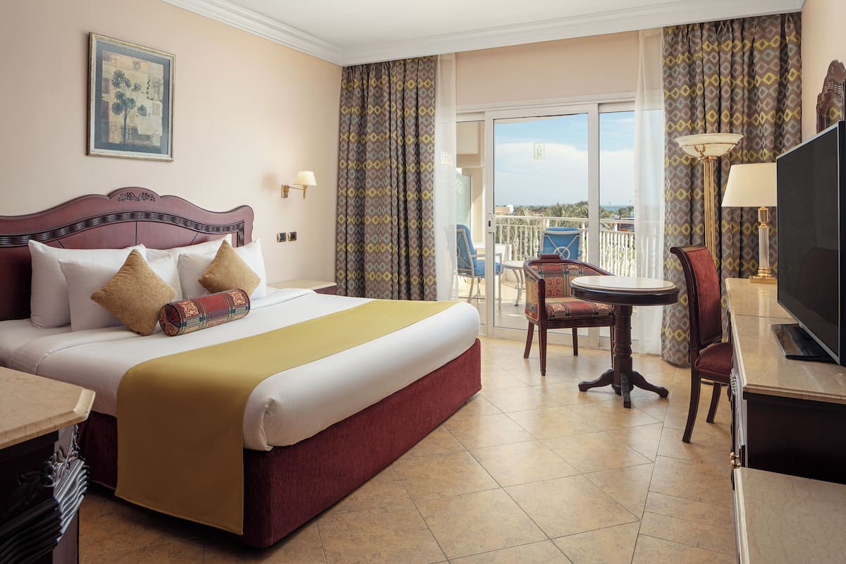 A hotel room with a large bed, a desk, a chair, a TV, and a balcony with a view.