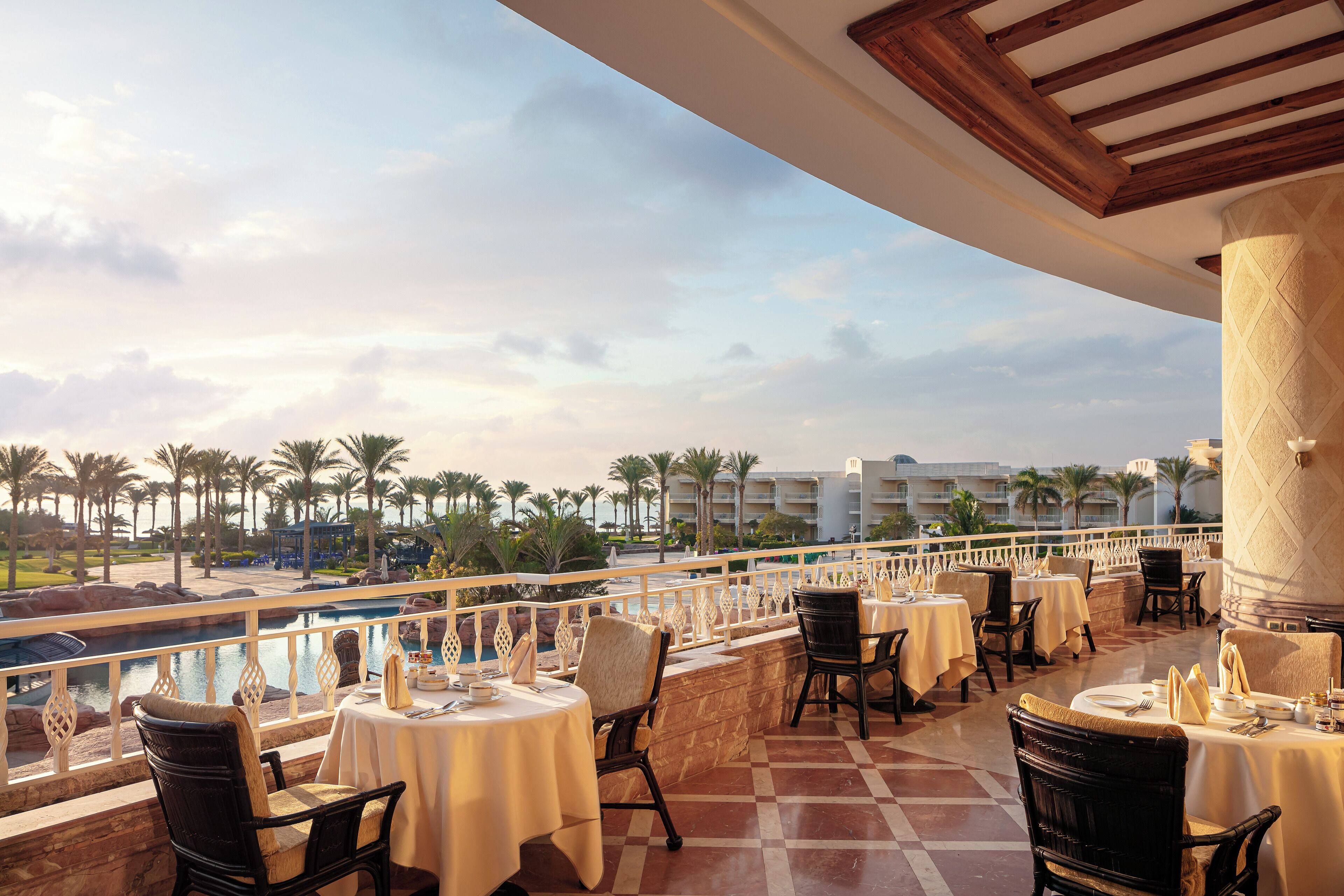 A terrace with tables set for dining, overlooking a pool and palm trees.