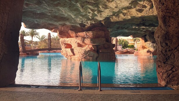 A pool area with a natural rock formation overhead, palm trees, and a bridge. - Palm Royale Resort - Soma Bay (Soma Bay)