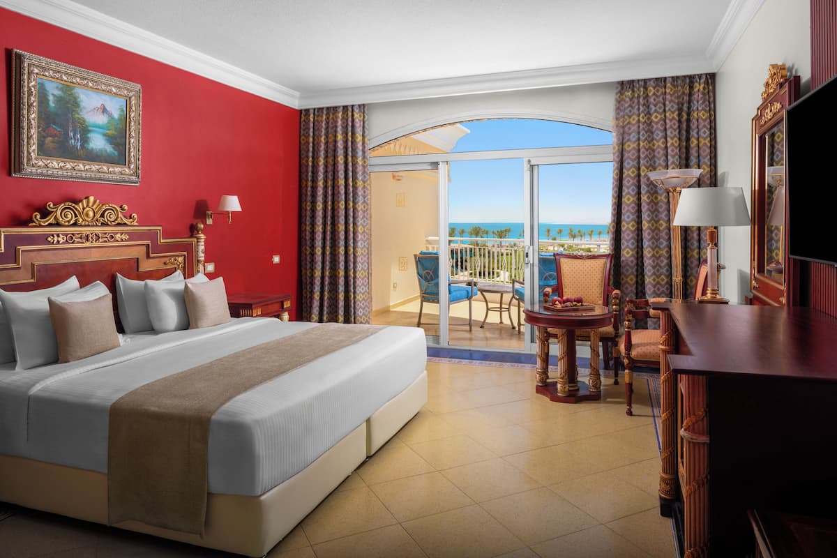 A hotel room with a large bed, a desk, a chair, a TV, and a balcony with a view of the ocean.