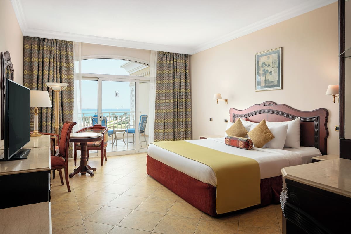 A hotel room with a large bed, a desk with a TV, a chair, and a balcony with a view.