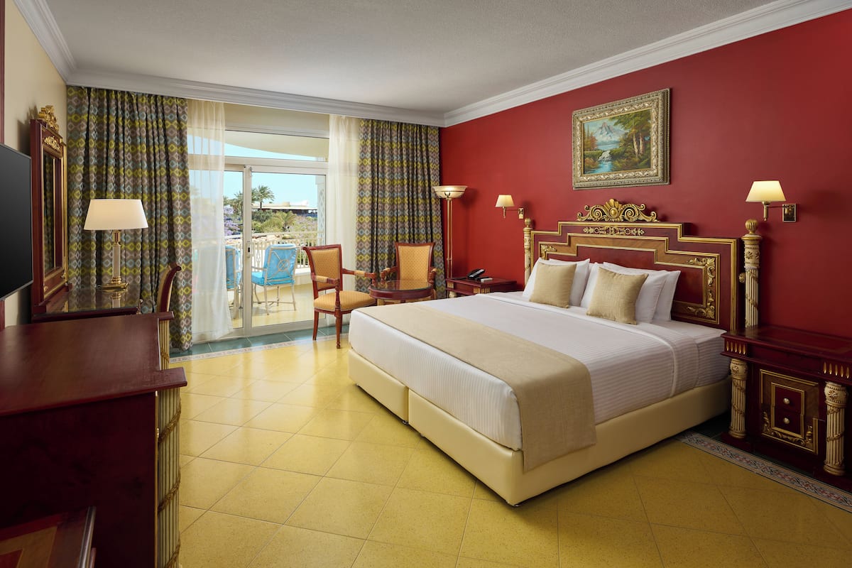 A hotel room with a large bed, a desk, a chair, and a balcony with a pool.