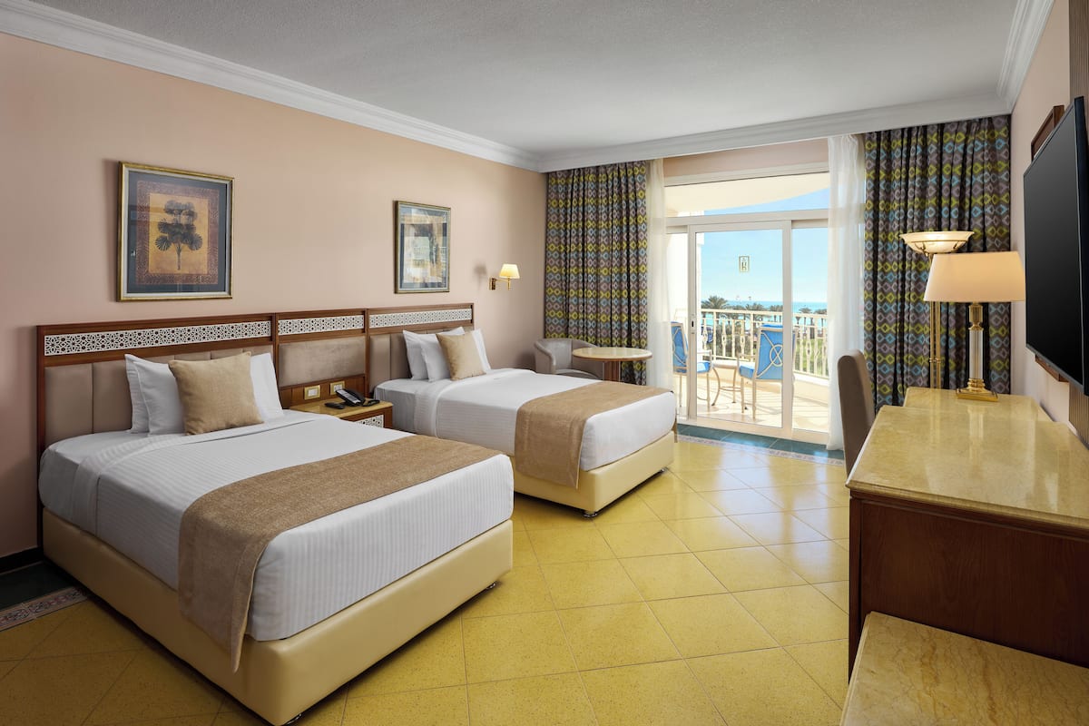 A hotel room with two beds, a desk, a chair, a TV, and a balcony with a view.