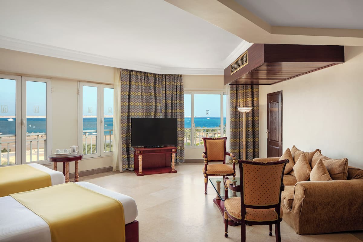 A hotel room with two beds, a TV, a sofa, and a balcony with a view of the sea.