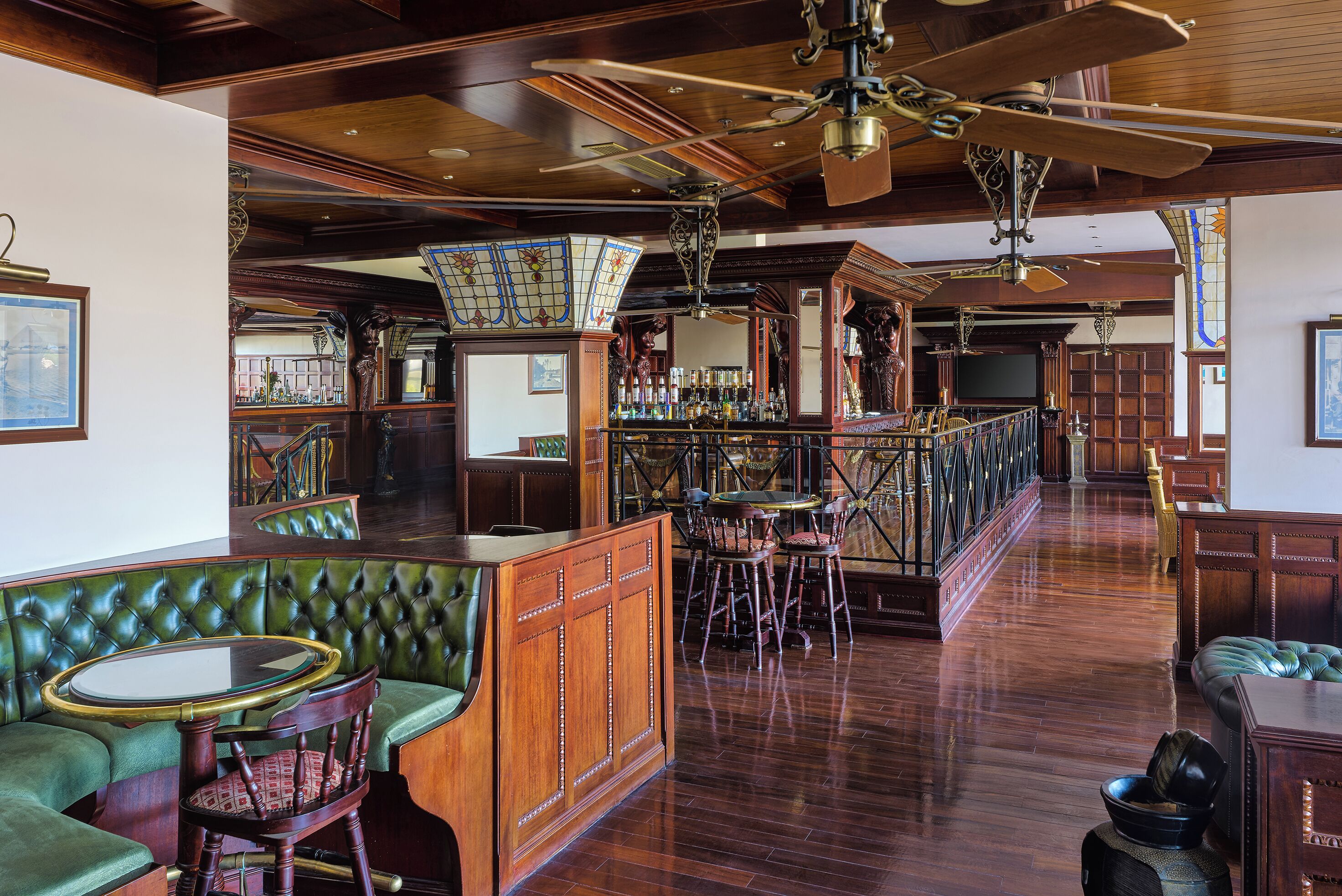 A bar area with a wooden bar counter, green upholstered seating, and a high ceiling with a fan.