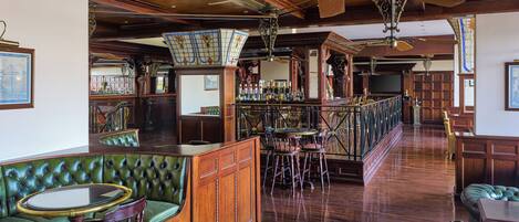 A bar area with a wooden bar counter, green upholstered seating, and a high ceiling with a fan.