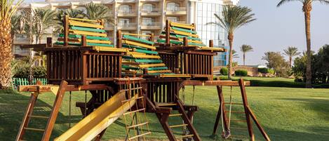 A playground with multiple slides and climbing structures in a grassy area with palm trees and a hotel in the background.