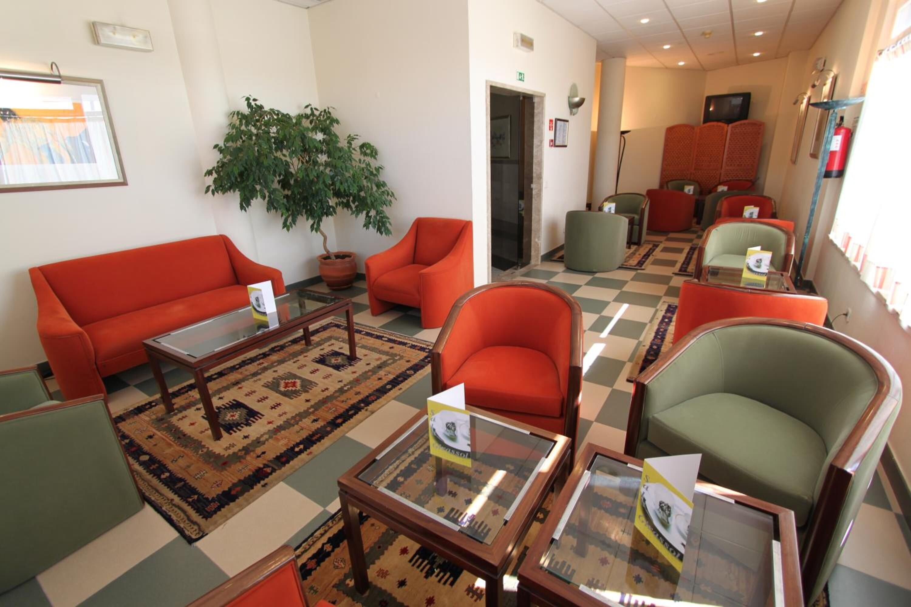 A lobby with a checkered floor, orange and green armchairs, a glass coffee table, and a potted plant.