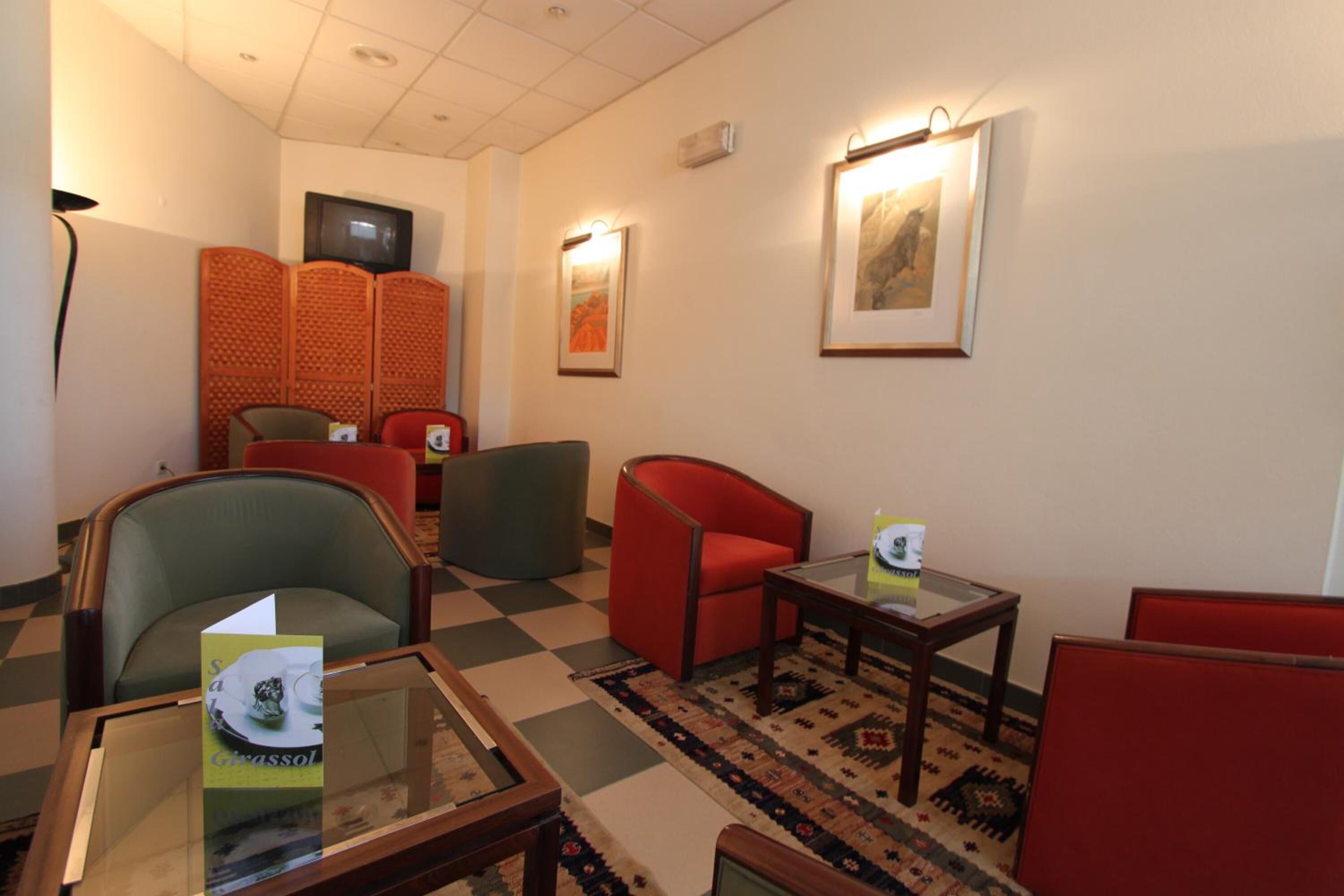 A hotel lobby with a checkered floor, red and green chairs, a glass-top table with a brochure, a television on the wall, and framed artwork.